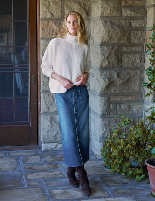A woman stands on a stone porch wearing a cream ribbed turtleneck sweater, a long blue denim skirt, and dark brown suede boots., view 4