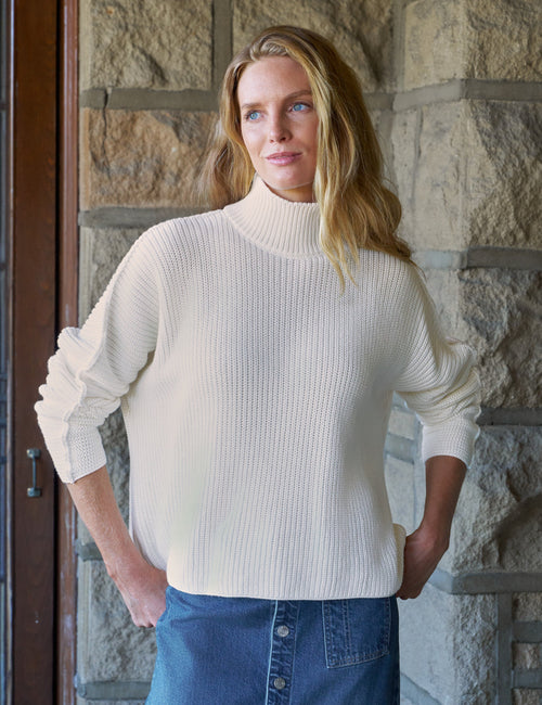 A woman wearing a cream ribbed turtleneck sweater and a blue denim skirt, standing in front of a stone wall., front view
