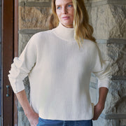 A woman wearing a cream ribbed turtleneck sweater and a blue denim skirt, standing in front of a stone wall.
