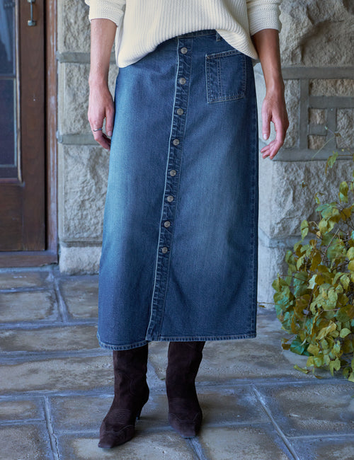 A person wearing a blue denim maxi skirt with a front button placket and a single front patch pocket, paired with a cream ribbed sweater and dark brown ankle boots, standing on a stone patio., front view