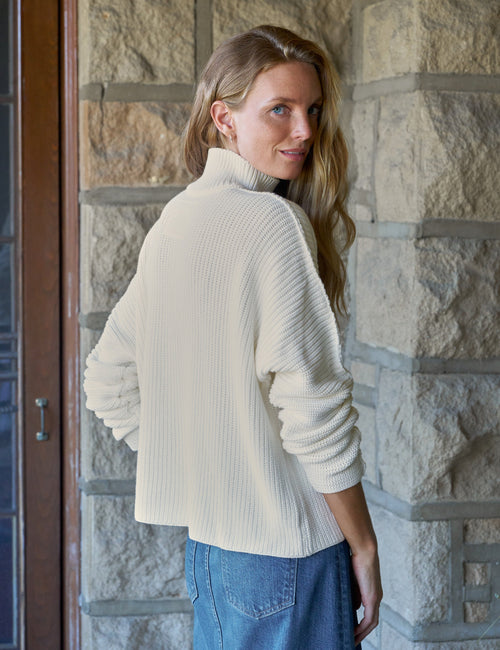 A woman with blonde hair is shown from the back wearing a cream ribbed turtleneck sweater and a blue denim skirt, standing in front of a stone wall., alternate view