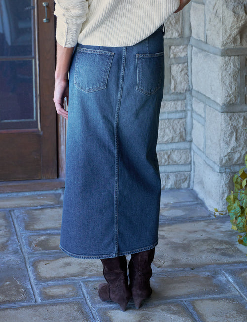 A person shown from the back wearing a maxi blue denim skirt with back patch pockets, a cream ribbed sweater, and dark brown boots, standing on a stone patio in front of a stone wall., alternate view
