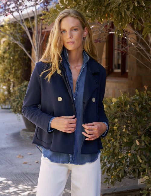 A woman wearing a navy blue double-breasted jacket over a blue denim shirt and white pants, standing outdoors in front of a building with greenery., front view