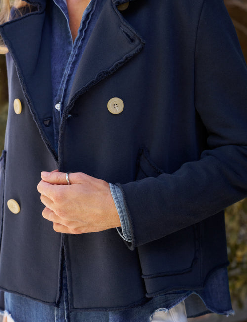A close-up of a person wearing a dark navy peacoat over a blue denim shirt and jeans, with the hands folded in front of the jacket., view 3
