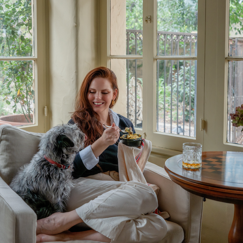 Woman sitting on a couch with a dog, eating from a bowl in a sunlit room.