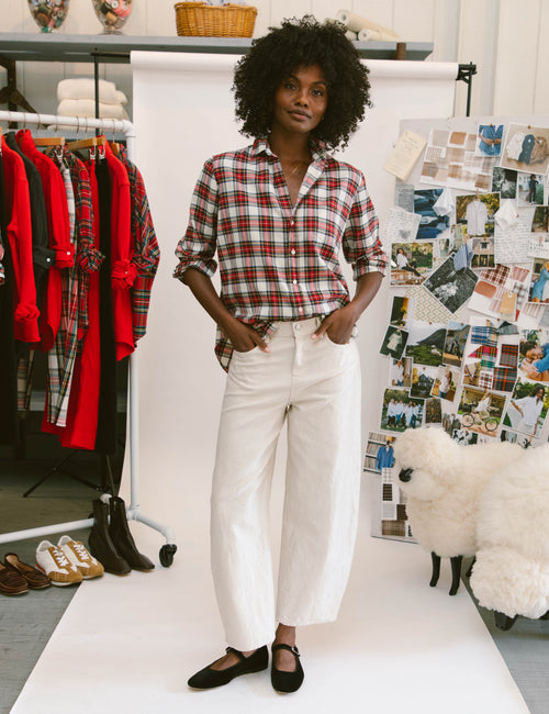 A model stands on a white runway in a studio setting, wearing a red and white plaid button-down shirt tucked into white cropped barrel-leg pants with black flats. To the left is a clothing rack with red and plaid garments, and to the right is a collage board with fashion photos and a white sheep prop., view 4