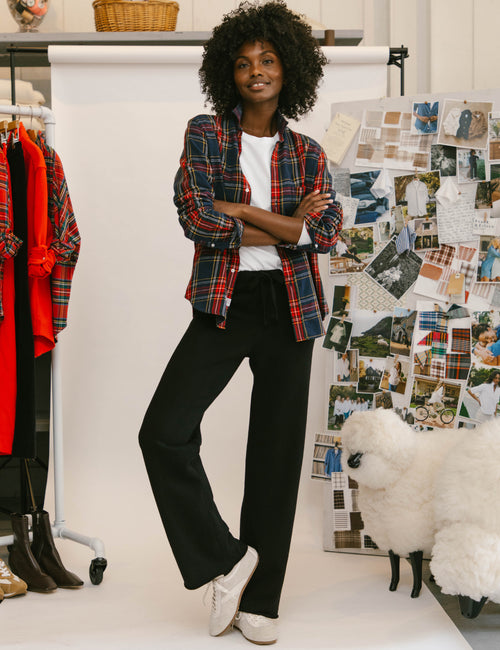 A woman stands in a studio setting wearing a red and blue plaid button-up shirt over a white top, black wide-leg pants, and white sneakers. A clothing rack with red plaid garments is visible on the left, and a collage board with fashion photos is on the right., view 6
