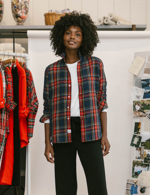 A woman with curly black hair stands in a boutique wearing a red, navy, and green plaid button-down shirt over a white t-shirt and black pants. To her left, a clothing rack holds red and black garments, and to her right, a white wall is decorated with fashion pins and notes., view 5