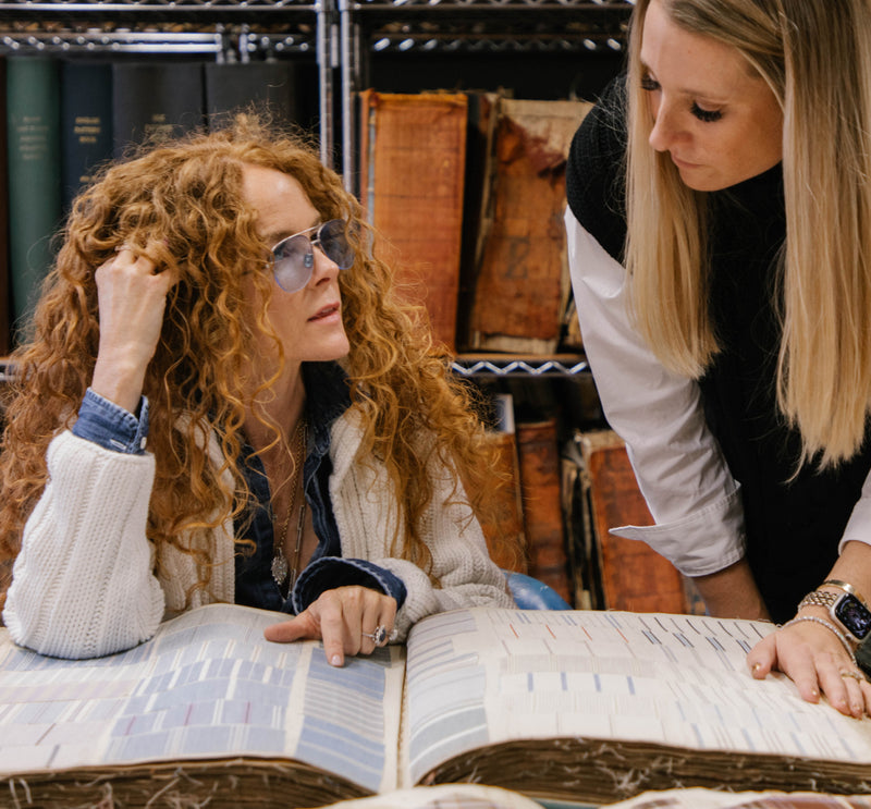 Two women examining an open large archival book or ledger in a study, with shelves of old books in the background.