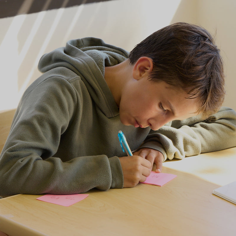 Young boy writing on pink paper with a blue pen on a wooden table.