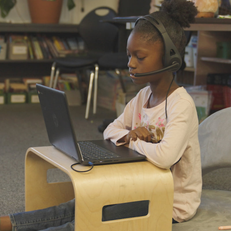 Child sitting on a stool using a laptop with a headset in a classroom setting
