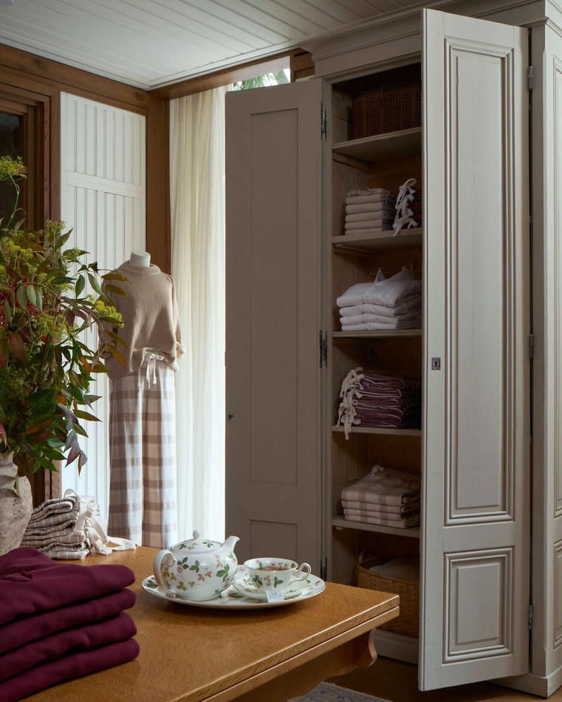 Open wardrobe with folded clothes in a room with a wooden table and teacup.