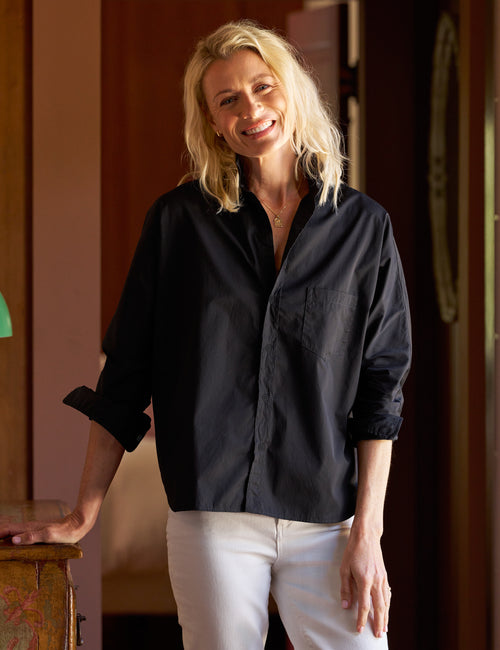 A smiling blonde woman wearing a loose black button-up shirt with rolled sleeves and white pants, standing indoors against a warm brown wooden background., view 5