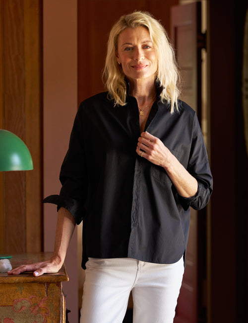 A woman with blonde hair wearing a black button-down shirt and white pants, standing in a warm-toned indoor setting., front view