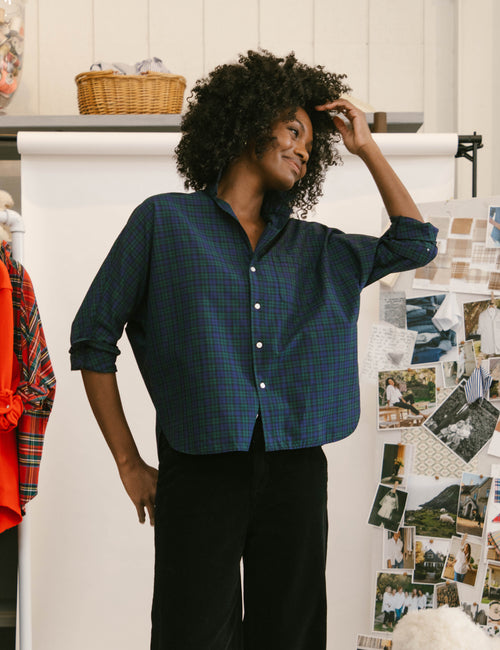 A woman with curly black hair wearing a dark green and navy plaid button-down shirt and black pants, standing in a room with a whiteboard and photos on a wall., front view