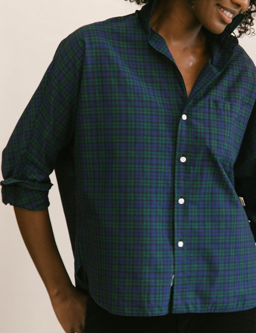 A woman wearing a dark green and blue plaid button-up shirt with white buttons, shown from the chest down against a plain light background., alternate view