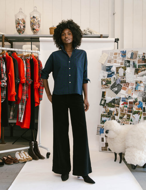 A model stands in a creative studio setting wearing a dark blue textured button-up shirt and black wide-leg trousers. To the left is a clothing rack with red and plaid shirts, and to the right is a white sheep prop and a board covered in fashion photos., view 4