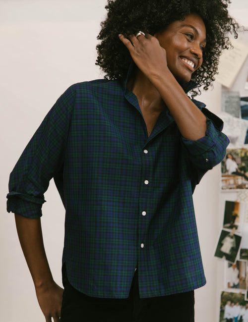 A woman with curly black hair smiling while wearing a dark green and black plaid button-down shirt with rolled-up sleeves, posed against a light wall with pinned photos in the background., view 3