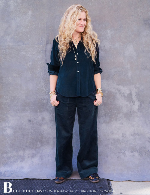A woman with long blonde hair stands against a textured gray wall wearing a dark navy button-up shirt and matching dark wide-leg pants. She is accessorized with gold bracelets and rings, and a photo credit for Beth Hutchens is visible at the bottom., view 5