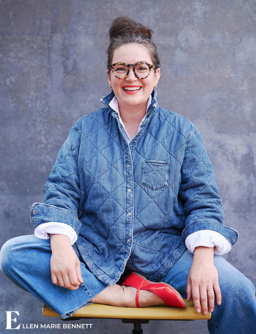 A smiling woman with glasses sits cross-legged on a stool against a textured gray wall, wearing a quilted blue denim jacket over a white collared shirt, blue jeans, and red pointed shoes. The Ellen Marie Bennett logo appears in the bottom left corner., view 7