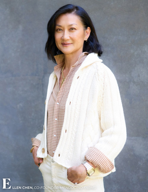 A woman with short dark hair stands against a textured gray wall wearing a cream knit cardigan over a brown and white gingham button-up shirt, paired with cream pants. A white logo watermark for Ellen Chen is visible in the bottom-left corner., view 6