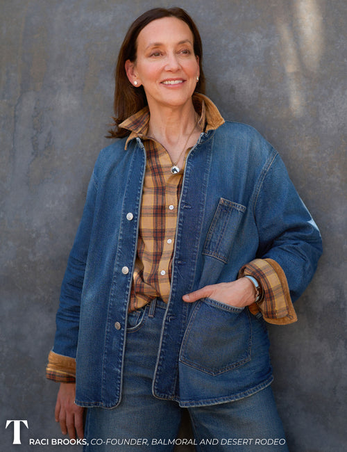 A woman wearing a blue denim jacket with tan lining over a brown and tan plaid shirt, standing against a textured gray wall. A white logo for 'Traci Brooks, Co-Founder, Balmoral and Desert Rodeo' is visible in the bottom left corner., view 4