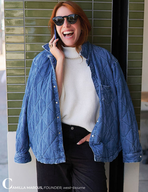 A woman wearing a blue diamond-quilted button-up jacket over a white ribbed turtleneck sweater and black pants, standing in front of a green tiled wall and holding a phone to her ear., view 8