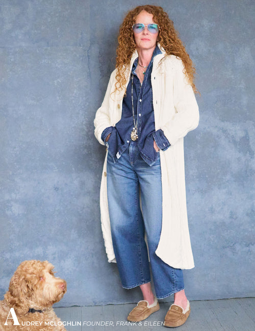 A woman stands against a textured blue wall wearing a long cream cardigan over a blue denim shirt and jeans, with tan loafers and sunglasses. A small dog is visible in the lower left corner., view 5