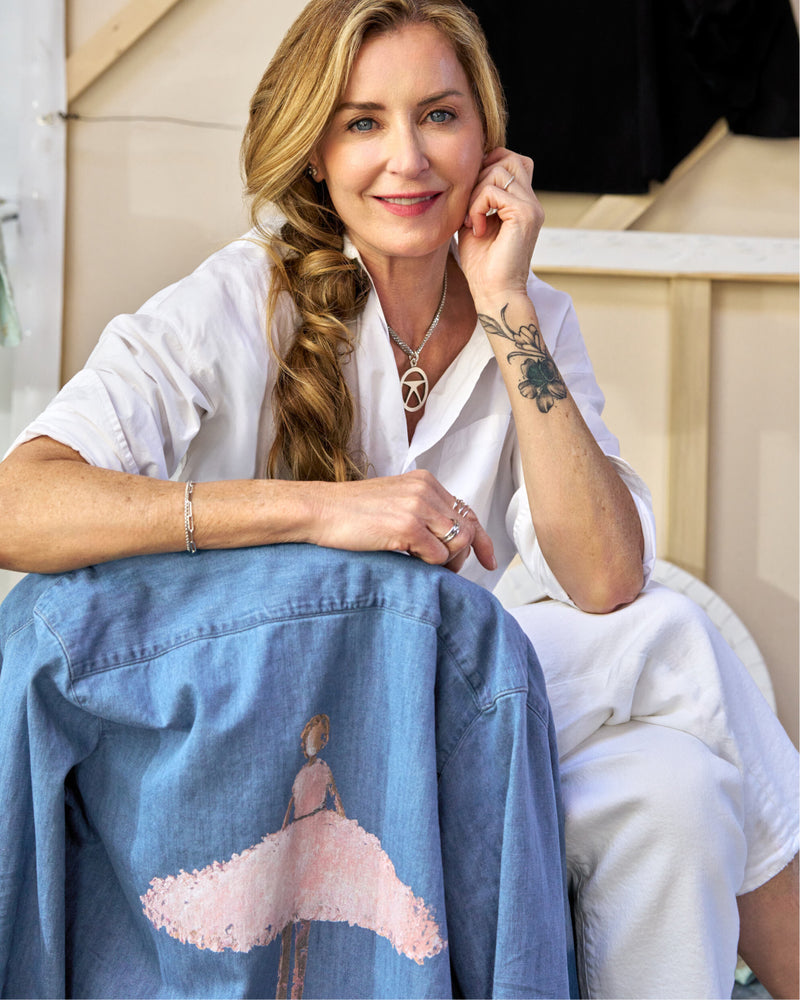 Woman wearing a white shirt and blue denim jacket with a design, sitting outdoors.