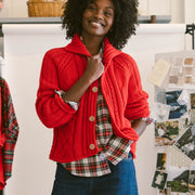 A woman stands in a styled indoor fashion setting wearing a bright red cable-knit button-up jacket over a red and black plaid shirt, loose dark blue wide-leg jeans, and black lace-up boots. A clothing rack with red plaid garments, a white sheep prop, and a collage board are visible in the background.