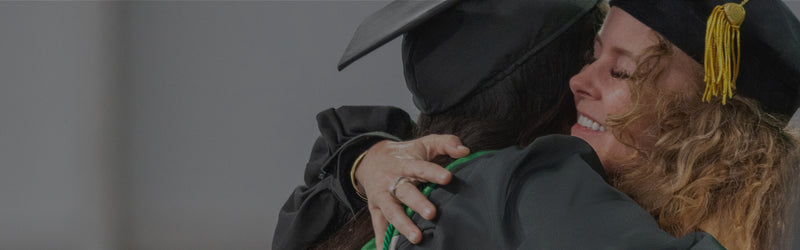 Two people in graduation caps and gowns embracing each other against a gray background
