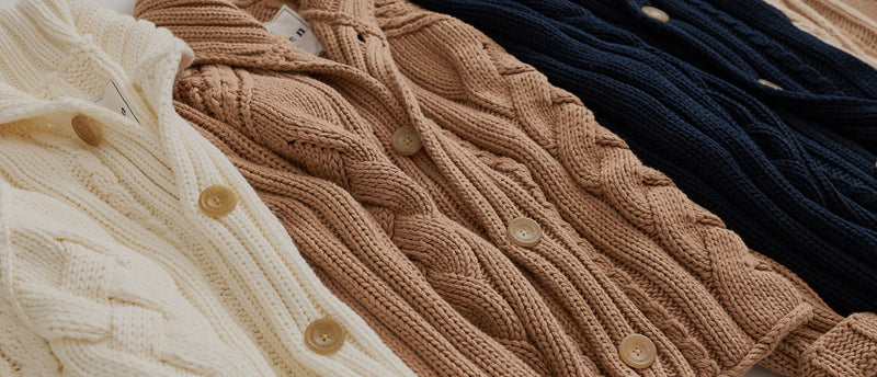 Three cable-knit cardigans in cream, tan, and navy blue, arranged diagonally with their buttons visible.